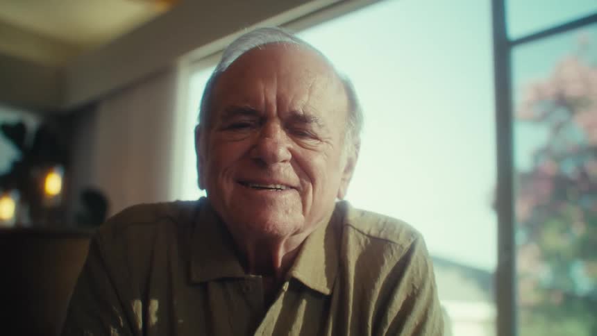 The video features a closeup of an elderly man with receding gray hair and darkrimmed glasses wearing a collared shirt he smiles warmly at the camera occasionally adjusting or removing his glasses with a bright sunlit room visible in the background