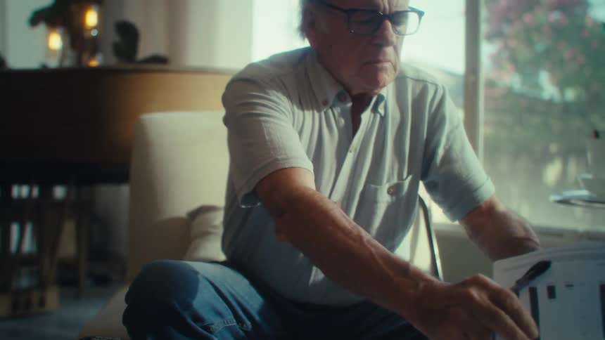 An older adult man wearing glasses and a light colored shirt sits at a table and uses a pen to write on documents then reviews other papers and a tablet with a grand piano and a large window looking out at trees in the background