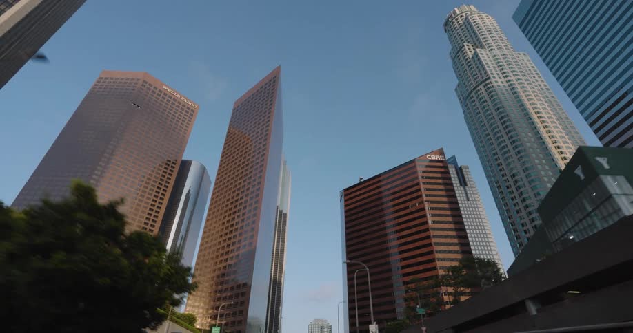 The video shows an upward view of a cluster of towering skyscrapers with various architectural styles and reflective facades reaching towards a clear blue sky