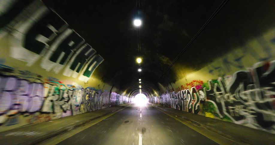 The video begins inside a long curved road tunnel with bright graffiti on its walls and overhead lights illuminating the path then moves forward to exit the tunnel into a city street with tall buildings and trees