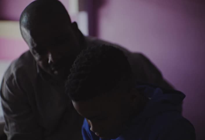 A young male child in a blue hoodie sits looking down while an adult male figure leans over him partially silhouetted against a light purple wall