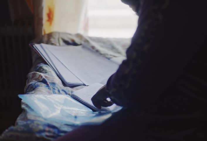 A silhouetted person sits in front of a window looking down at an open book or notebook with light coming from the window illuminating the pages