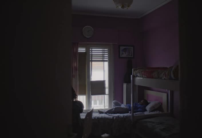 Two kids are inside a bedroomone lies on the bed another sits on a chair beside it and a girl walks in and out of the room