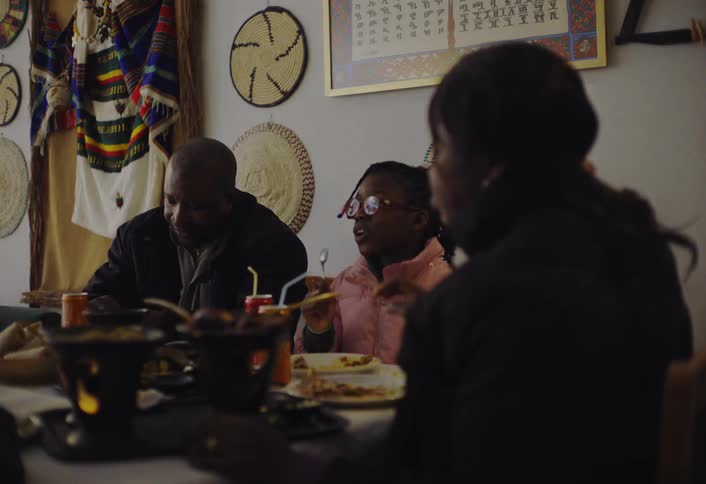 A family gathered around a table in a restaurant or dining room eating from various dishes while conversing with decorative items on the wall in the background