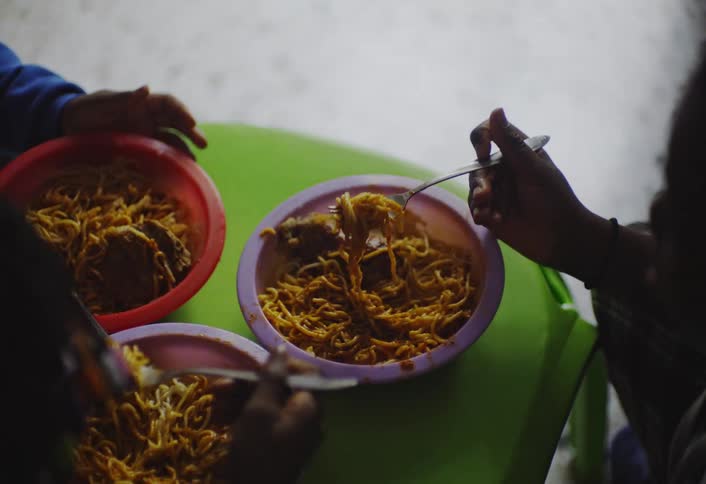 Three young children sit around a green table each eating noodles from a colorful bowl with a fork or spoon