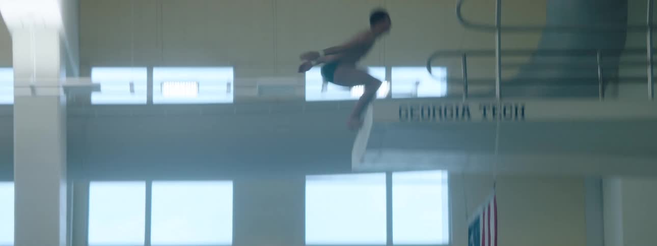 A male swimmer wearing only swim trunks jumps off a diving platform into the pool