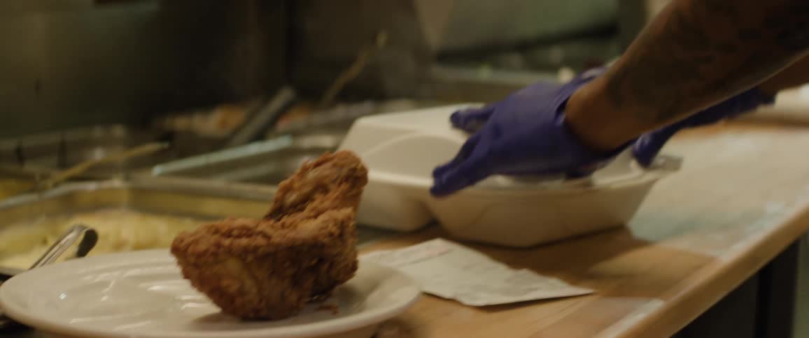 The video shows a closeup of a person wearing blue gloves carefully packing pieces of fried chicken into a white takeout container another piece of fried chicken rests on a white plate in the foreground next to a paper receipt