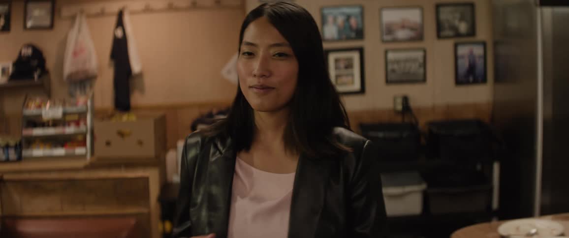 A woman with dark hair and a black jacket gazes thoughtfully to her right positioned in the foreground of a softly lit indoor setting with another person visible in the background