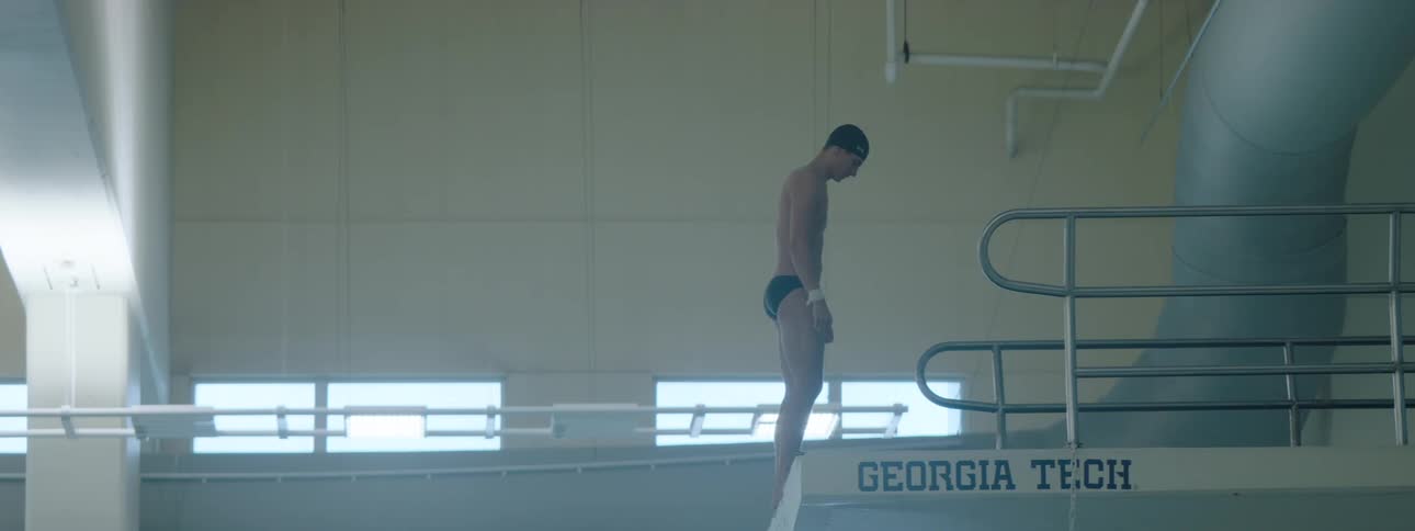 The video shows a male diver in a black swimsuit walking towards the edge of a diving board labeled georgia tech at a swimming pool he stops at the edge and takes a moment to prepare before facing forward
