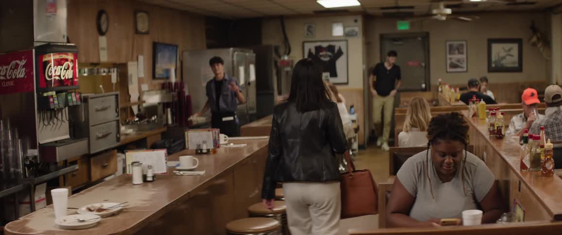 The video shows the interior of a casual diner or restaurant with wooden booths and tables and a long counter with stools customers are seated at various tables and some staff members are visible behind the counter and moving through the space
