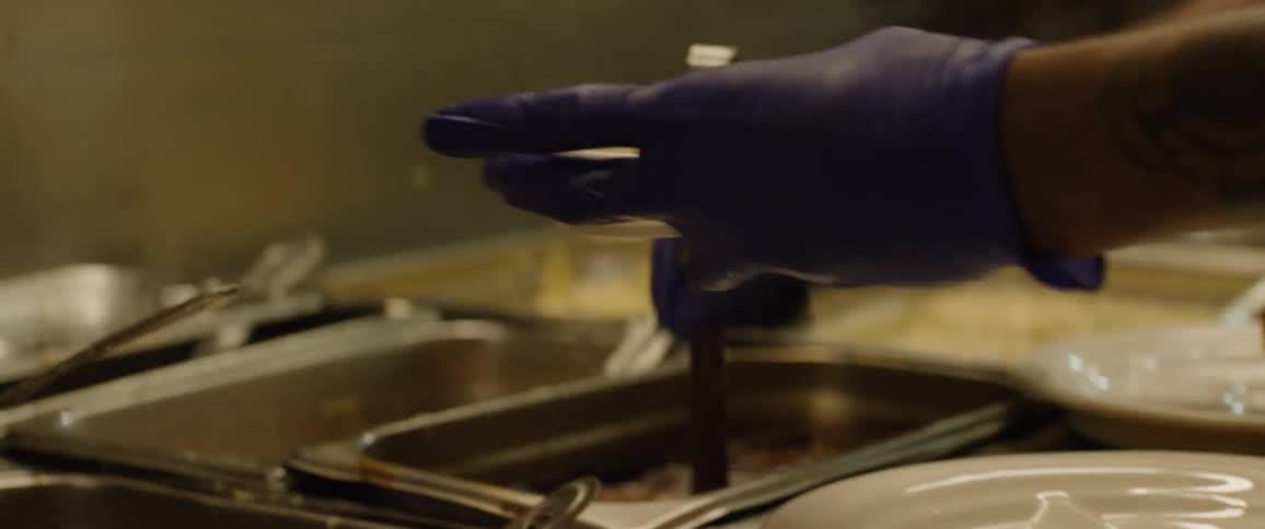 A cook wearing a blue glove places a piece of fried chicken on a white plate then uses tongs to add mashed potatoes from a serving tray and finally scoops gravy onto the potatoes in a kitchen setting