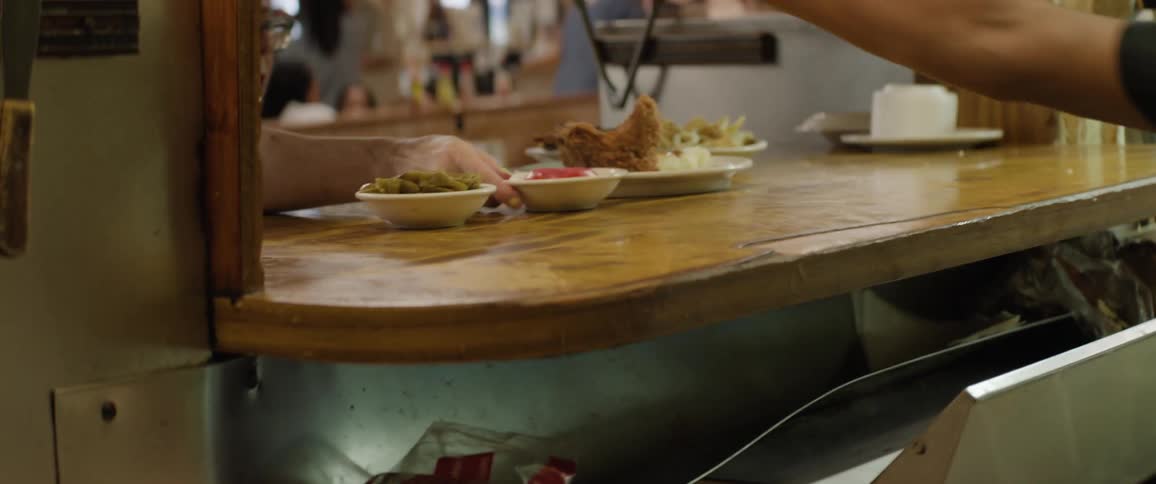 A cook wearing blue gloves slides a plate of fried chicken and mashed potatoes across a wooden counter in a busy kitchen while taking new orders