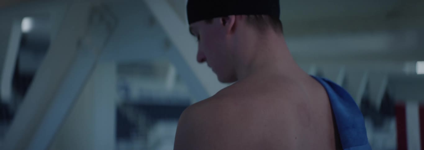 A tall male swimmer wearing a cap walks past an indoor pool