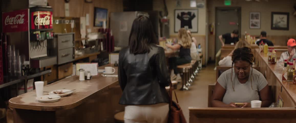 The video shows a busy diner interior with several patrons seated at booths and at a long counter a woman with dark hair seen from behind walks through the diner eventually turning to face the camera with a slight smile