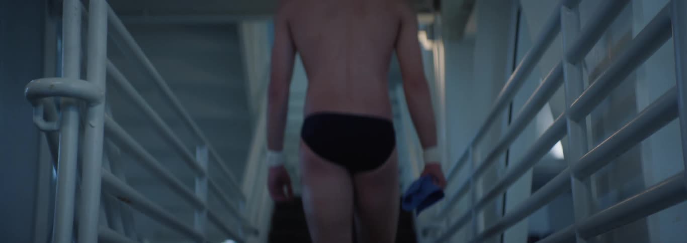 A young adult man wearing black swim briefs and a swim cap walks up a set of white stairs carrying a blue towel in what appears to be an indoor swimming facility