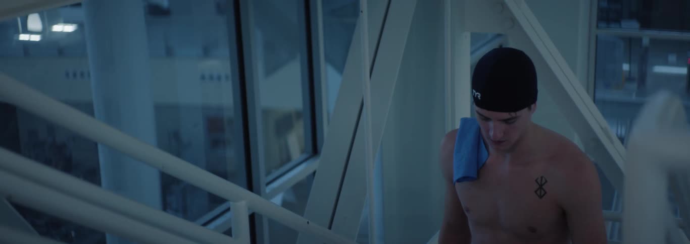 The video shows a swimmer seen from the chest up with wet hair and a black swim cap appearing to walk out of the pool area a blue towel is draped over his left shoulder