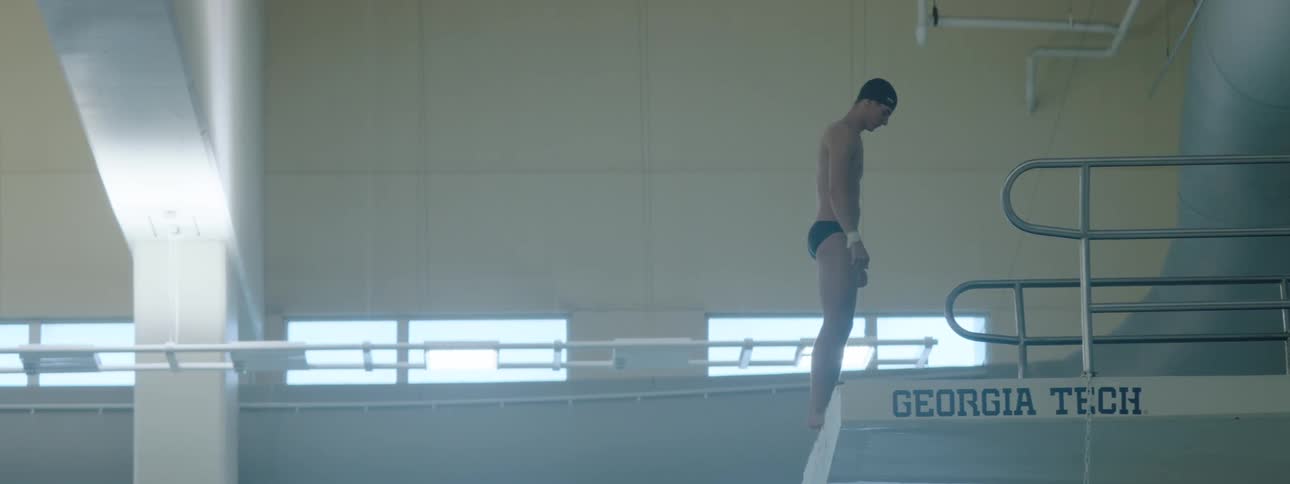A swimmer walks to an elevated diving board at midday wearing swimming trunks and posing ready to dive into the pool