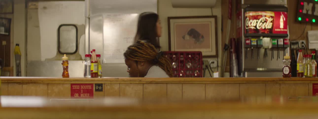A young adult female employee with dreadlocks is seen from behind a wooden counter in a diner kitchen bending down to retrieve something then rising as another young adult female employee walks by