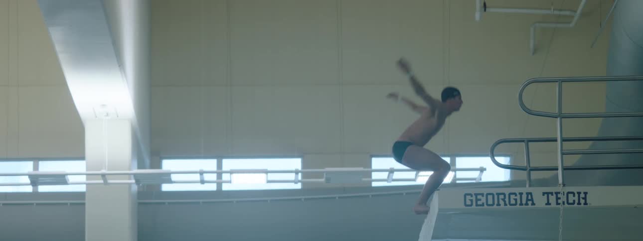 The video shows a male diver standing on a diving board with his back to the camera his arms extended upwards in preparation for a dive he then brings his arms down and begins to prepare for his dive