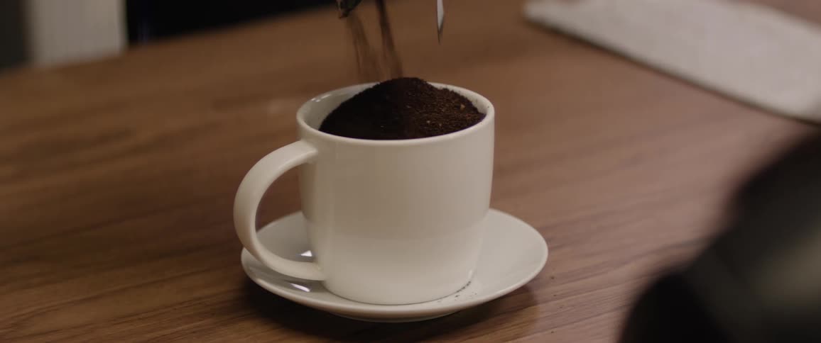 A white coffee mug on a matching saucer sits on a wooden table as dark coffee grounds are poured into it creating a small mound above the rim with a metal coffee pot in the background