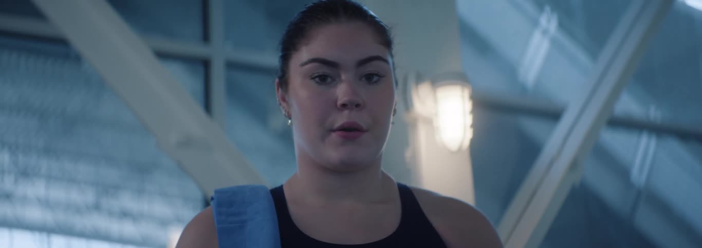 The video shows a female swimmer seen from the chest up with dark hair tied back and a blue towel draped over her shoulder she looks directly at the camera with a determined expression