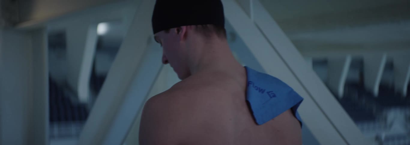 A close view of an american flag hanging in a large indoor stadium with empty blue seats then shifts to show a young adult man wearing a black swim cap and holding a blue towel walking past large structural beams