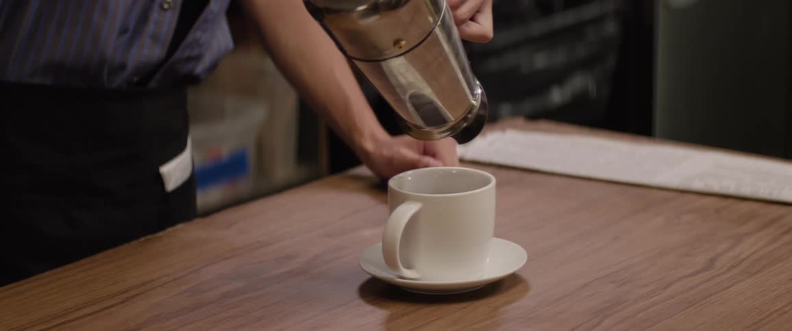 A white coffee mug sits on a wooden counter and a baristas hands are seen pouring a mound of dark coffee grounds from a metal coffee pot into the mug