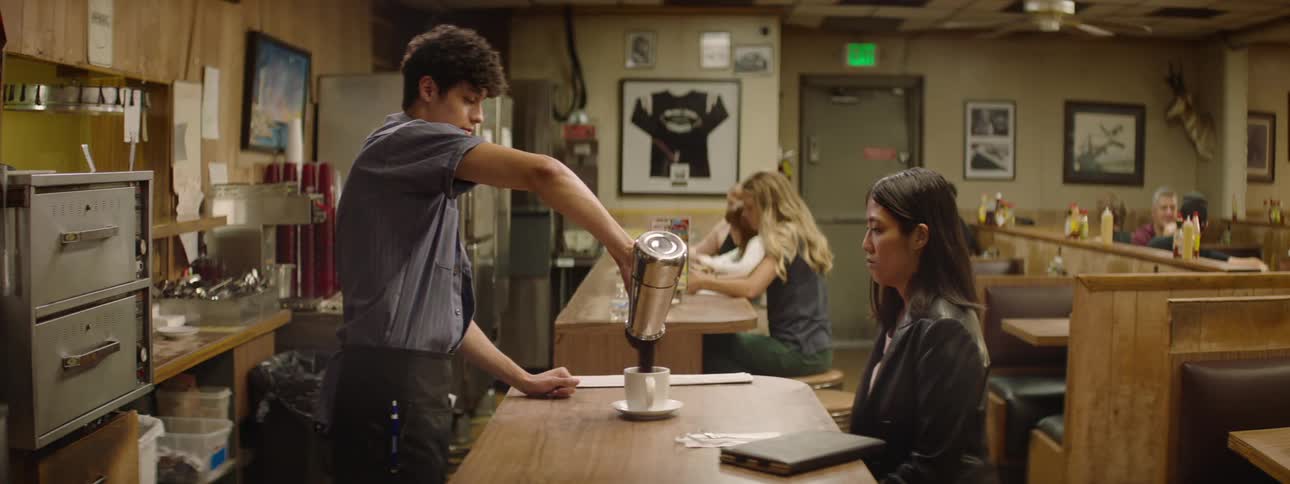 Man serving coffee from a pot to a woman sitting at a table in a coffee shop