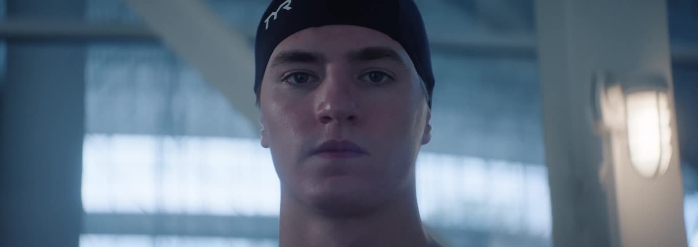 A young adult male swimmer wearing a black swim cap looks directly at the camera with a determined expression with light blue and white blurred structures in the background suggesting an indoor swimming pool