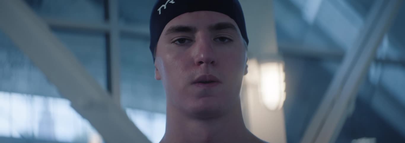 A young adult male swimmer wearing a black swim cap looks directly at the camera with a determined expression with light blue and white blurred structures in the background suggesting an indoor swimming pool