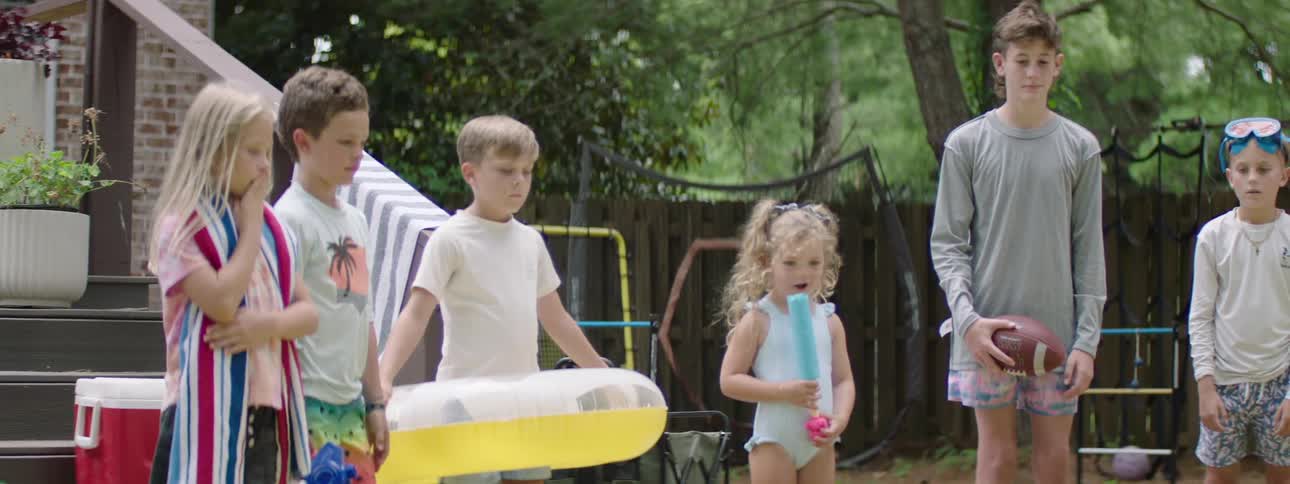 The video shows four young children standing in a grassy backyard likely preparing for water play as evidenced by a yellow and white inflatable float the girl on the far right is holding a blue toy and the other children appear to be in swim attire
