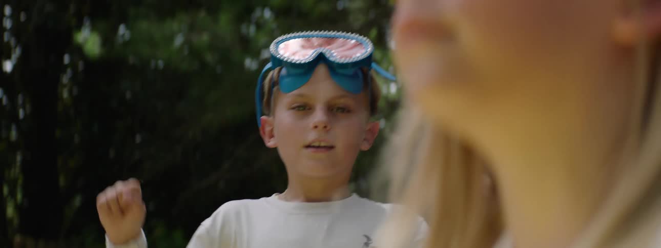 A young boy wearing blue swimming goggles on his forehead and a white long sleeved shirt looks towards the camera with a playful expression raising his fists in the air with blurred green trees in the background