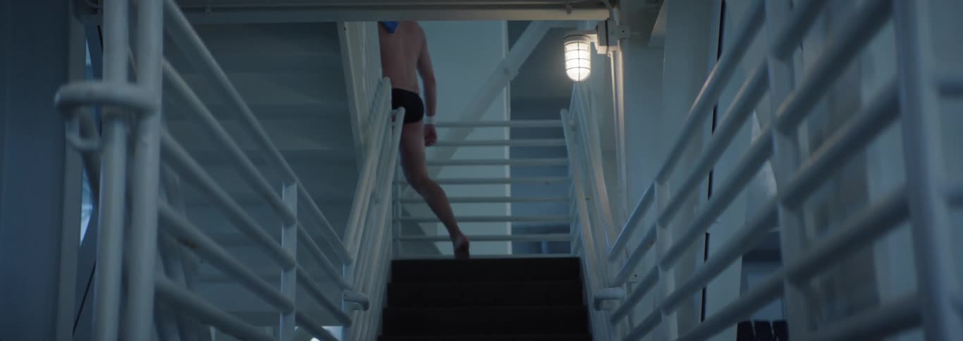 The video shows a male diver viewed from behind and wearing black swim briefs walking down a set of stairs towards a pool area the stairs have white railings and the scene is lit by overhead lights suggesting an indoor aquatic facility