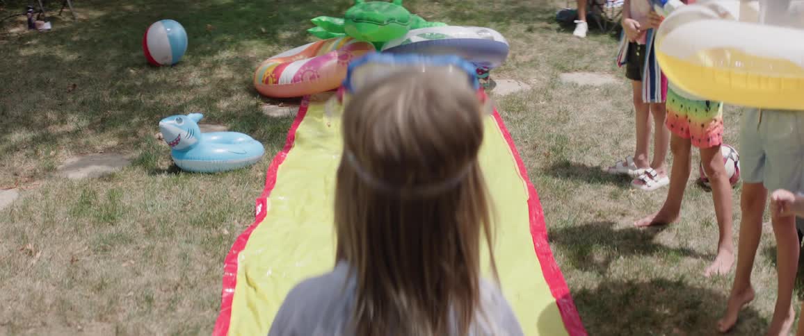 The video shows the back of a young boy wearing a white tshirt and blue and green patterned swim shorts he is standing on a green lawn with a yellow and red water slide laid out in front of him