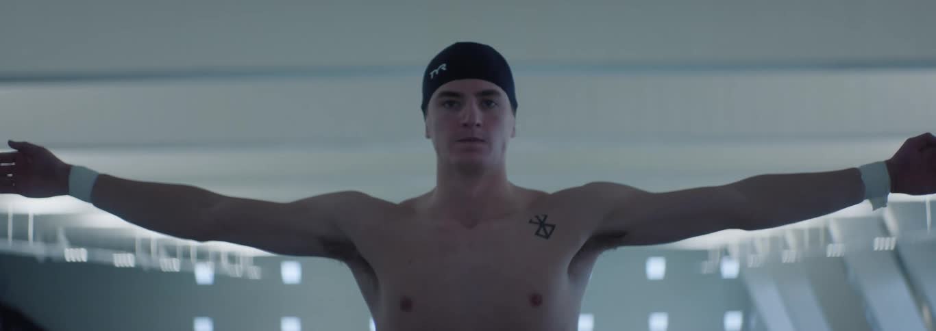 A young male swimmer wearing a swim cap stands shirtless in an indoor pool facility looking focused outstretching his arms