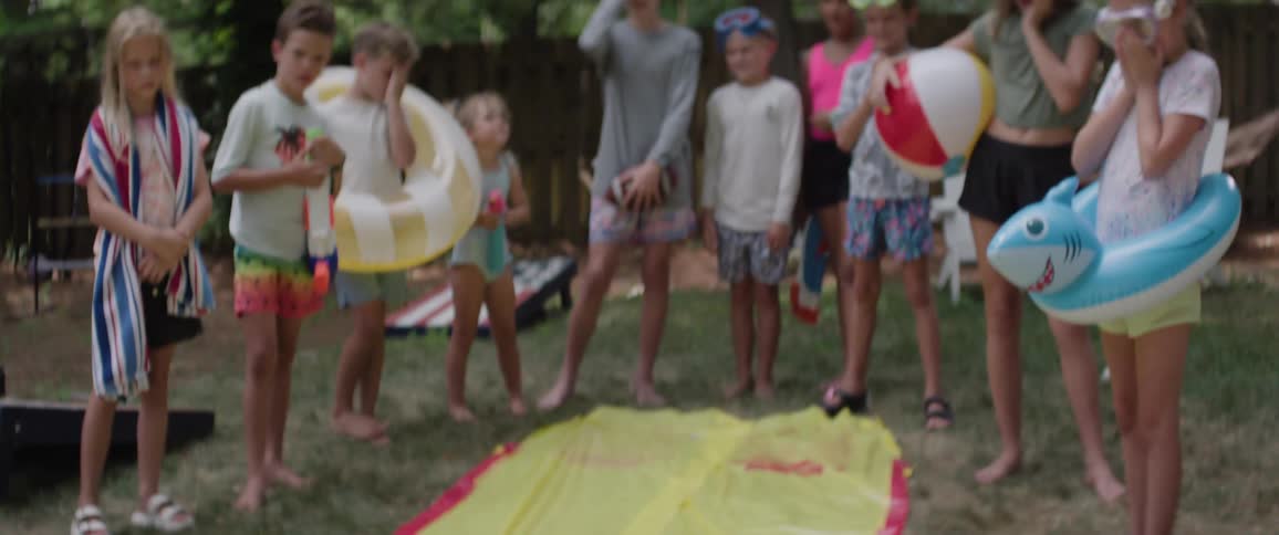 A young child in swim goggles glides facefirst on a yellow slipandslide cheered on by barefoot friends gathered on the grass
