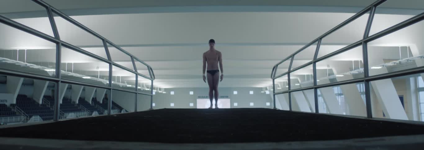 Man in swim trunks with arms outstretched doing an acrobatic jump into an indoor pool