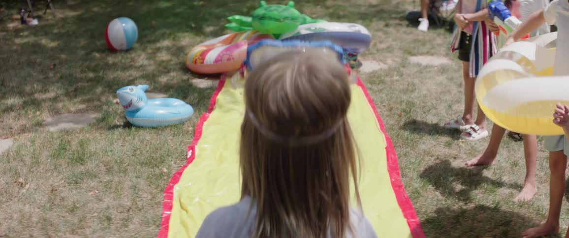 The video shows a childs legs and lower torso from behind standing on a green lawn with a yellow and red water slide laid out in front of them the child is wearing blue and green patterned swim shorts