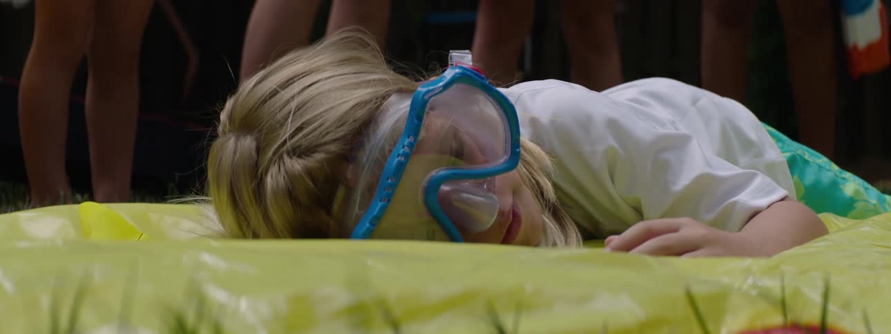 Child drops to their stomach on a yellow plastic bag on the grass in front of other people
