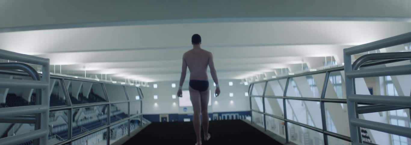A male athlete swimmer walks on a diving platform toward the edge wearing only swim trunks preparing to dive into the pool