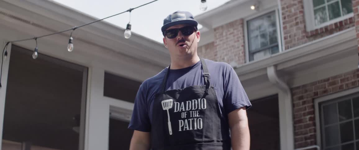 The video shows a man with a mustache and sunglasses wearing a baseball cap and a black apron that says daddio of the patio he is holding a metal grill spatula