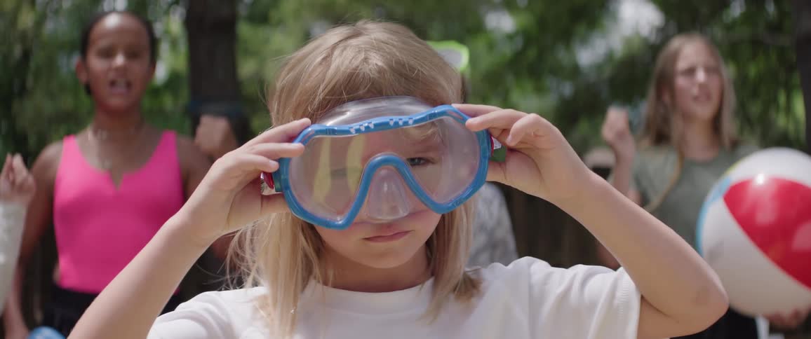 Little girl putting on swimming goggles and other people in the background cheering in a backyard