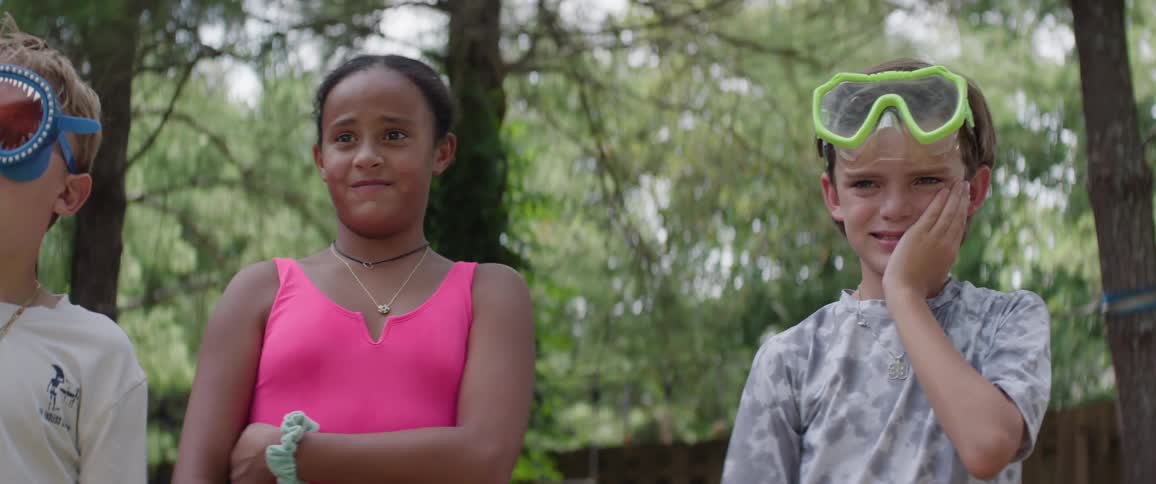 The video features a closeup of two children outdoors against a backdrop of green trees a girl in a pink top initially stands with a neutral expression while a boy next to her wearing green swimming goggles on his forehead holds his hand to his face