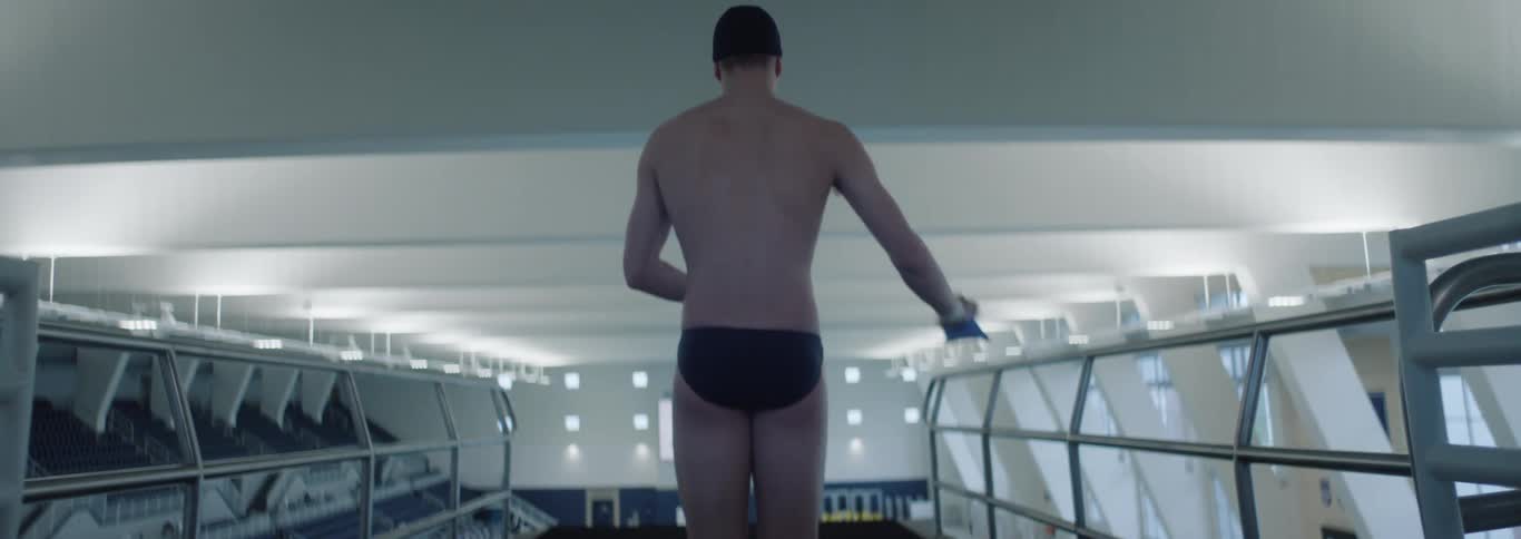 The video shows a male diver viewed from behind wearing black swim briefs and a swim cap with a blue athletic tape visible on his left shoulder he walks purposefully towards the edge of a diving board with rows of empty stadium seating visible on eit
