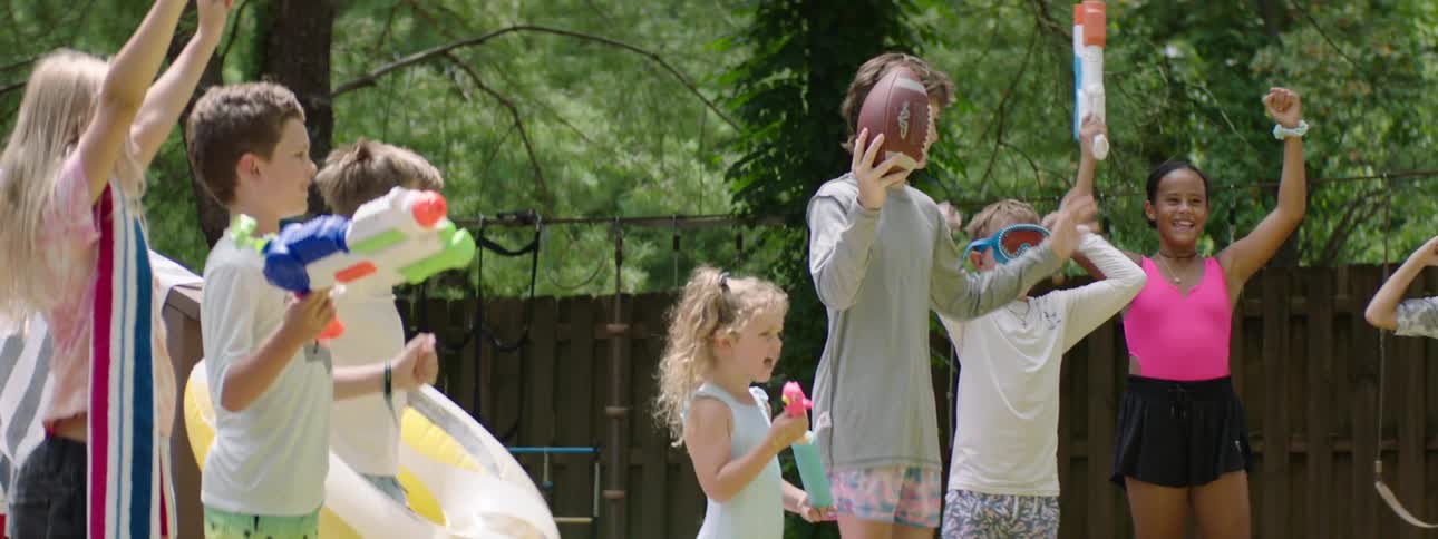 The video shows a group of children playing outdoors on a sunny day with several holding water guns and one boy holding a football they appear to be enjoying themselves smiling and raising their arms in excitement