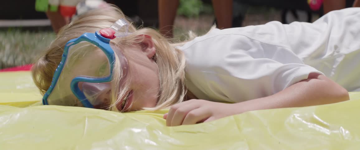 Little blonde girl with swimming goggles on her face falling on an inflatable balloon on the grass in the backyard