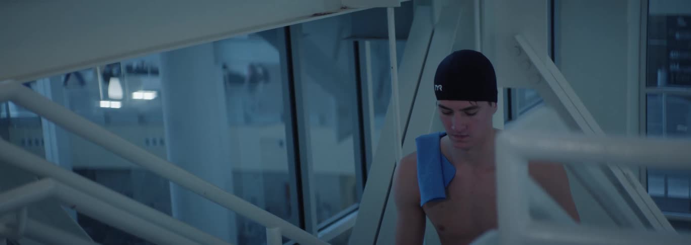 A swimmer wearing a black cap and blue towel walks through a sleek glassenclosed stairwell his expression focused and reflective in the cool modern setting