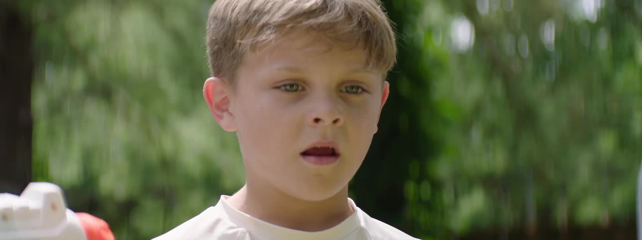 A young boy outdoors at midday surrounded by others looks shocked and surprised his mouth open as he covers his face with his hands