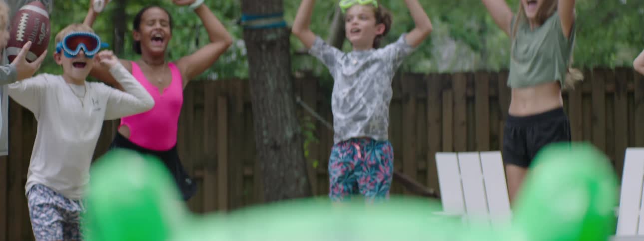 The video features a group of children and adults enjoying a backyard on a sunny day in the foreground a young girl with blonde hair and goggles looks towards the camera while another child with dark hair in a pink top smiles in the background