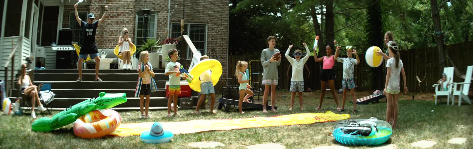 The video captures a lively backyard scene with children and adults engaged in outdoor fun several children are gathered around a yellow and blue water slide some holding inflatable toys and a football suggesting active play